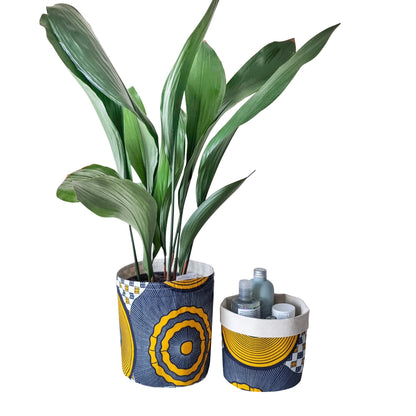 Two Osime Home Plant and Storage Pots. One on the right has a leafy plant stored inside it with the pot top rolled up. Next to it is the same plant and storage pot with the top slightly rolled down. Toiletries are stored Inside.